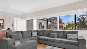 Ein modernes Wohnzimmer mit einem grauen Ecksofa, einem kleinen Hocker und einem großen Fenster mit Blick auf die Nachbarschaft.