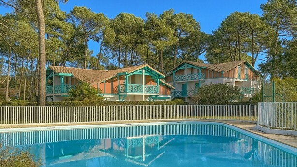 A swimming pool with a white fence, surrounded by two-story buildings with green balconies and a clear blue sky. - T2 Apartment mezzanine with terrace and pets allowed (Soustons)