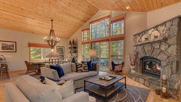 A cozy living room with a stone fireplace, large windows, and a wooden ceiling.