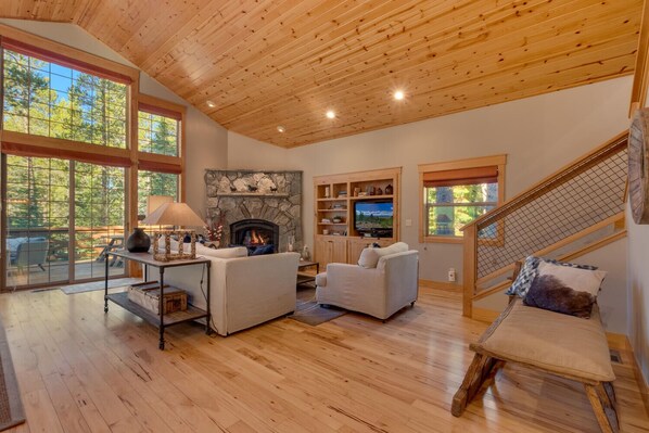 A spacious living room with a stone fireplace, wooden ceiling, and large windows offering a view of the outdoors.