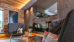 A modern interior with a wooden ceiling, a seating area with a cushioned chair and sofa, a small wooden table, and a large window offering a view. - Stadtvilla Schladming Boutiquehotel (Schladming)