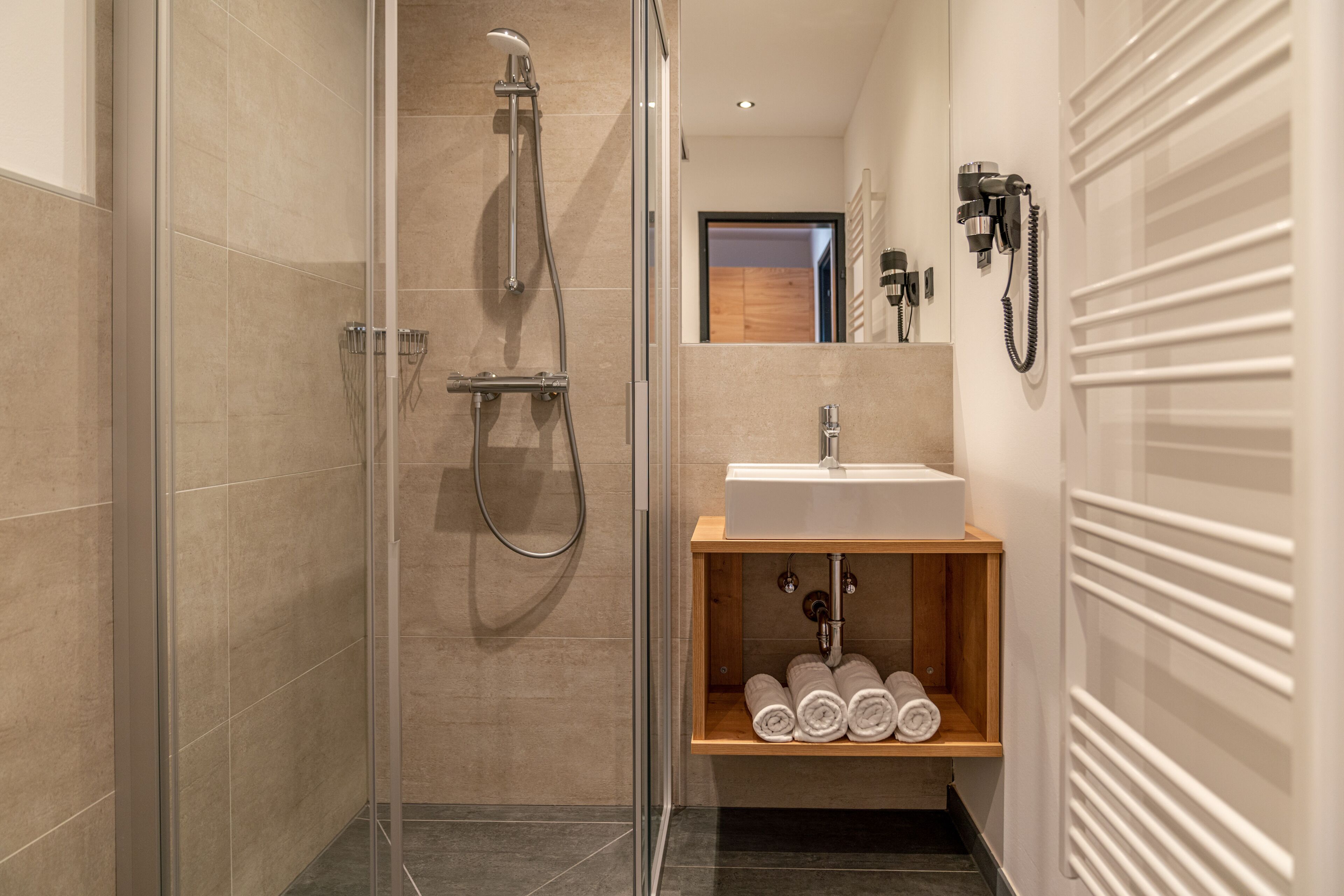 A modern bathroom with a glass-enclosed shower, a wooden shelf with towels, a sink, and a mirror.