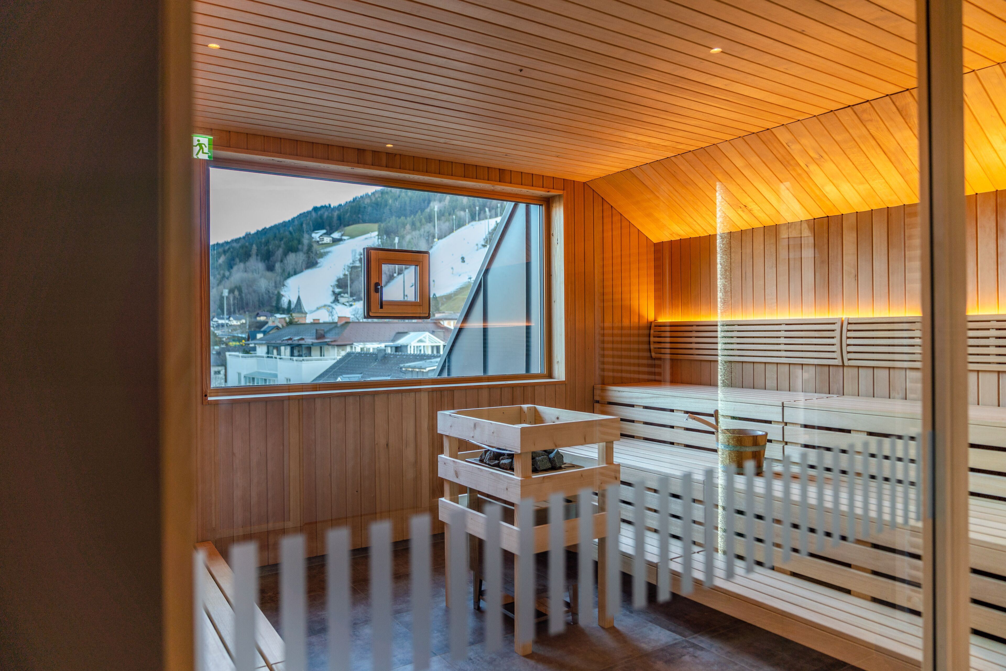 Un sauna con listones de madera, un banco de madera, una mesita de madera y una ventana con vistas a un paisaje nevado.
