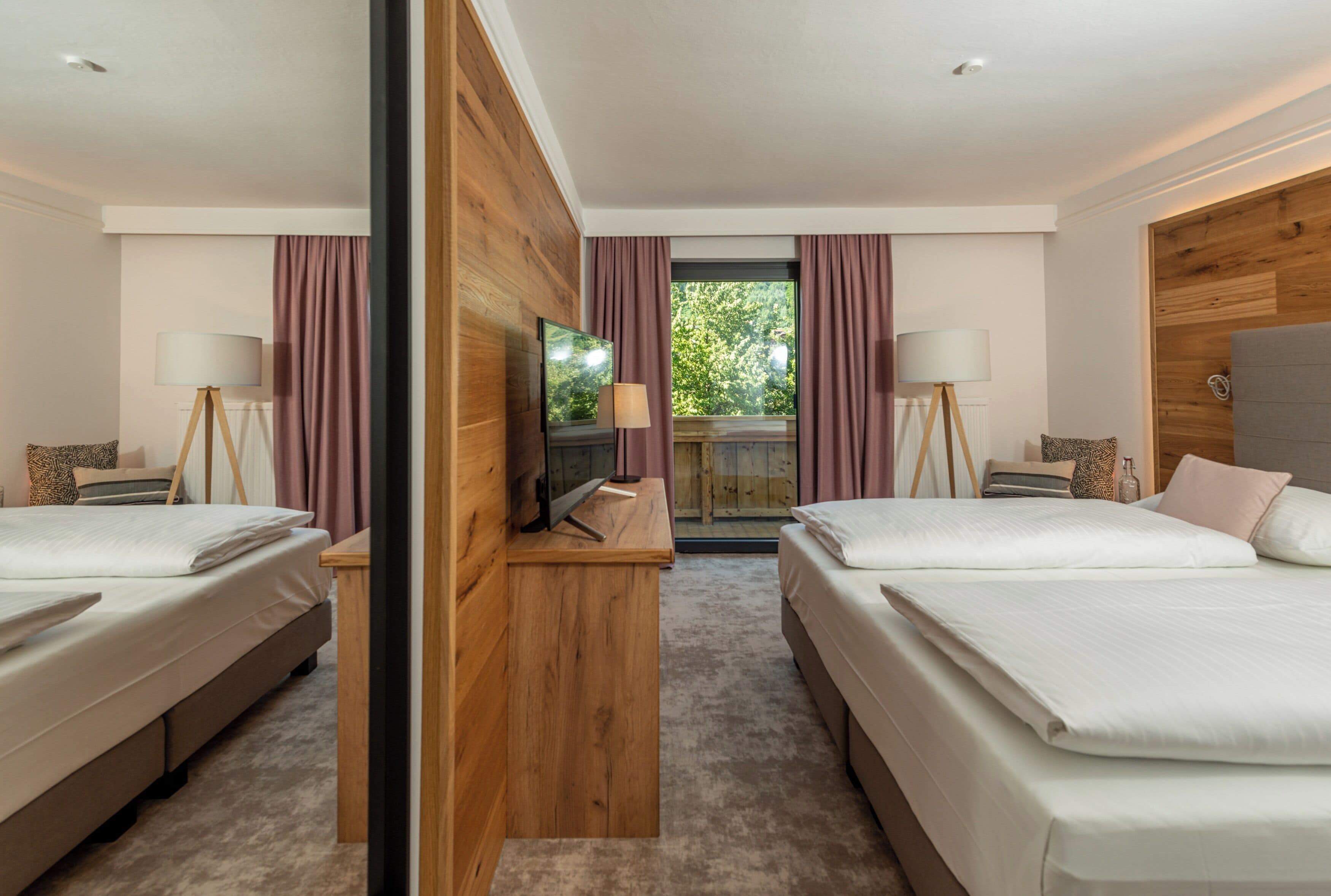 Habitación de hotel con dos camas, un escritorio de madera, televisión y balcón con cortinas.