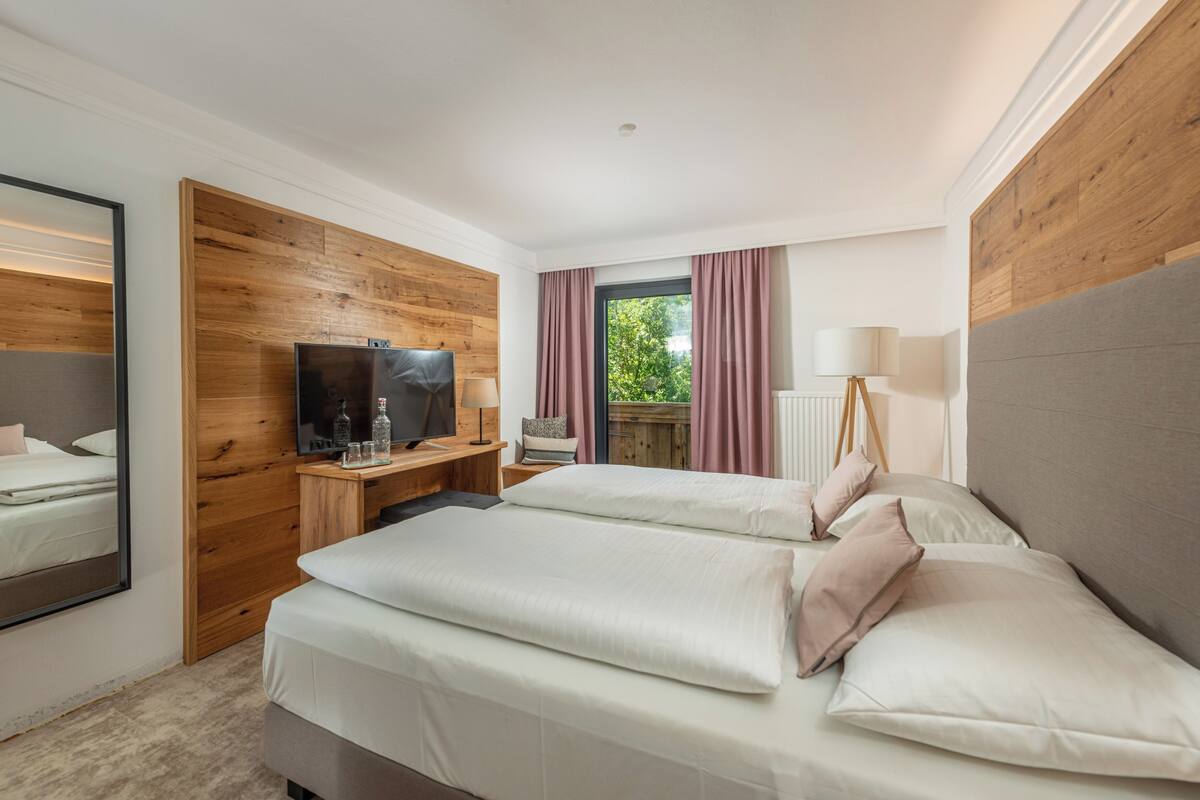 A hotel room with a wooden accent wall, a TV, a desk with a lamp, two beds, and a window with curtains.