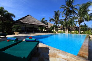 A swimming pool with lounge chairs, a thatched-roof building, and palm trees. - Villa Bunga Melati - Beachfront Vacation Villa in Bali (Dencarik)
