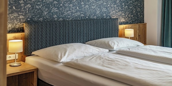 Ein Doppelbett mit weißen Leinentüchern, ein dunkel strukturiertes Kopfstück und zwei Nachttischlampen auf Holznächtigungstischen.
