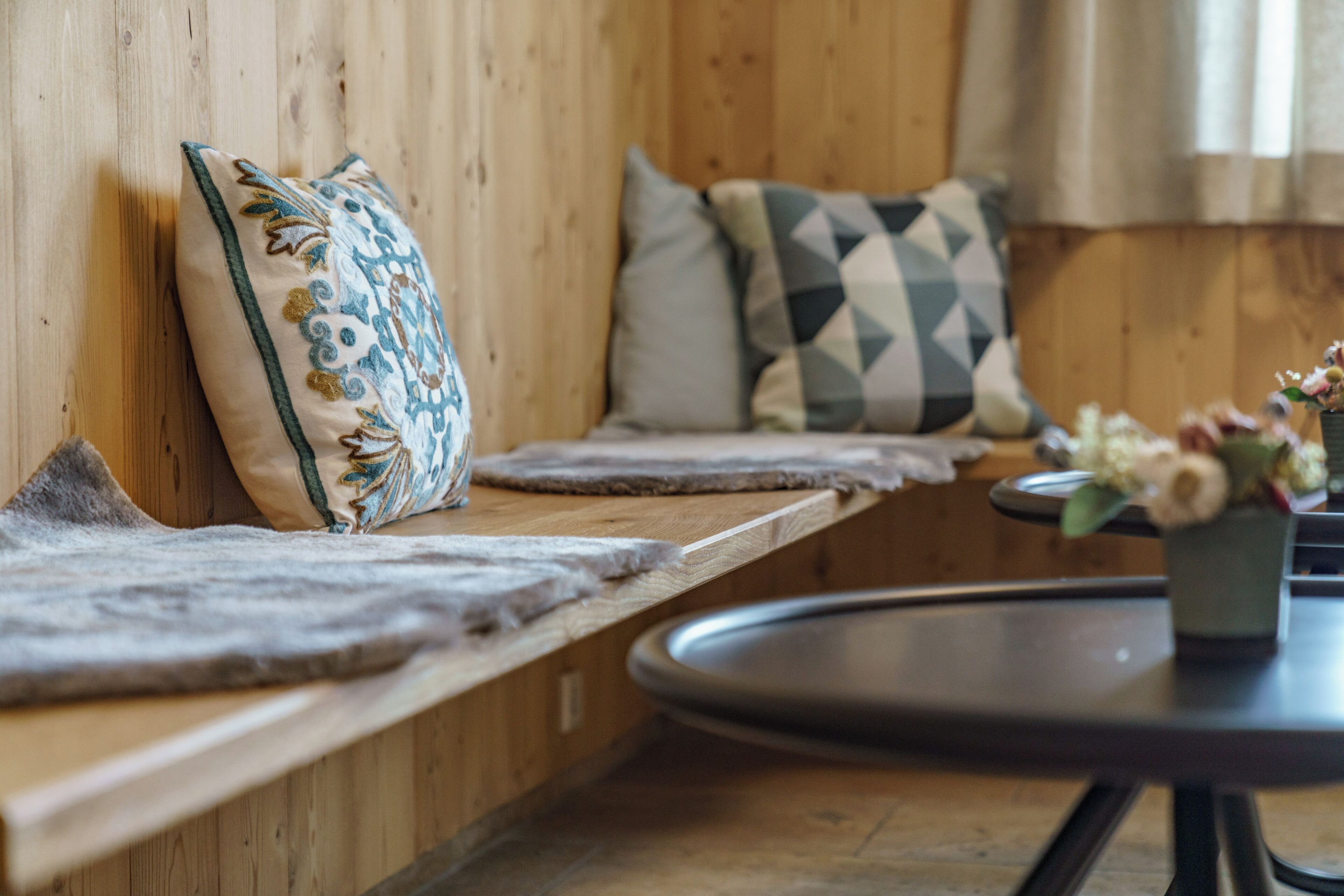 A wooden bench with cushions, a round table with a flower vase, and a wooden wall.
