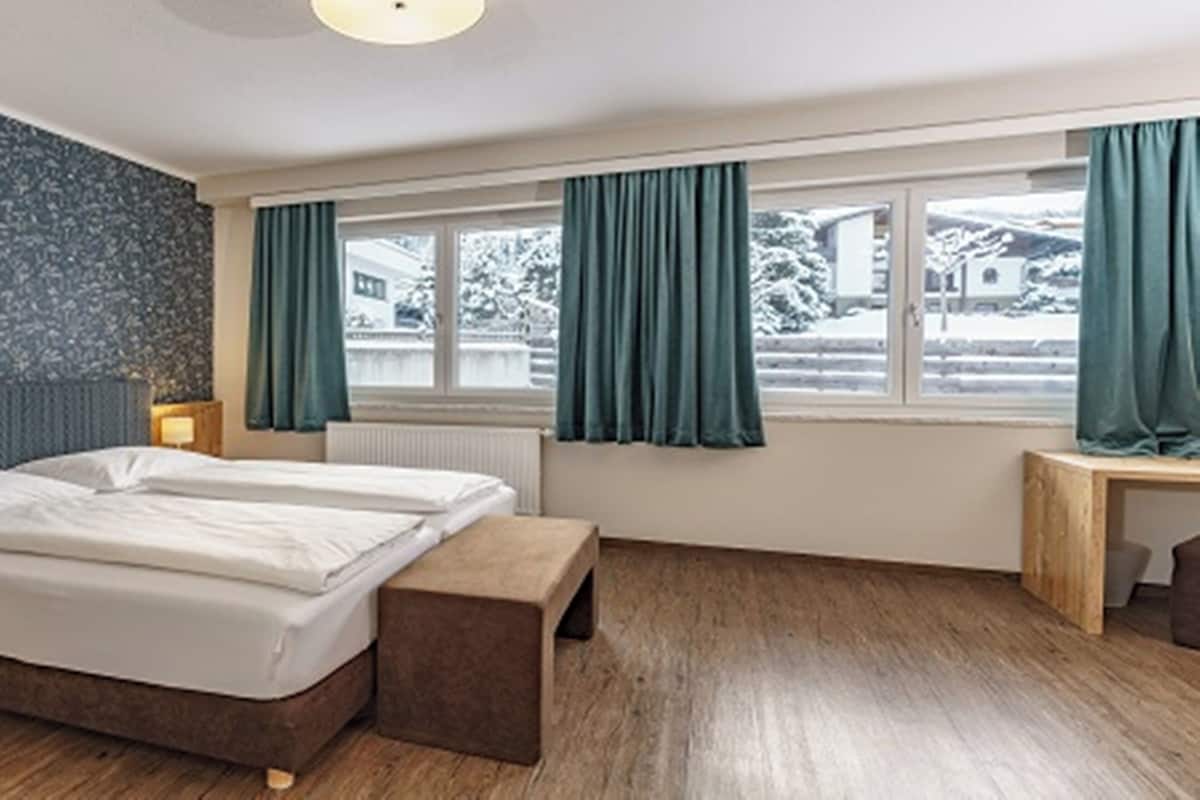 Une chambre d’hôtel avec un grand lit, un banc en bois, une table de chevet avec une lampe, une fenêtre avec des rideaux et une vue sur un paysage enneigé.