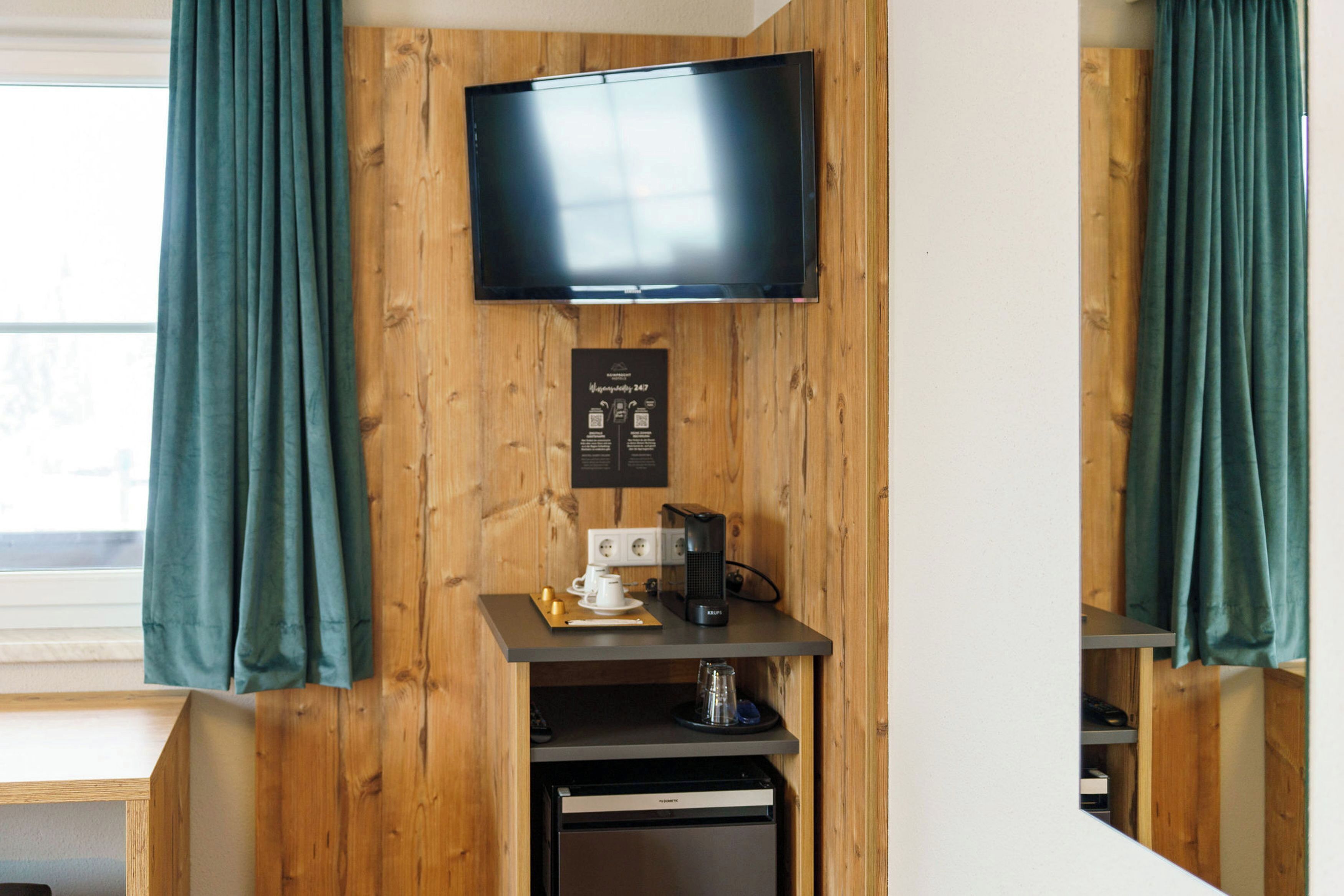 A room with a flat-screen TV mounted on a wooden wall, a coffee machine, and a small refrigerator.