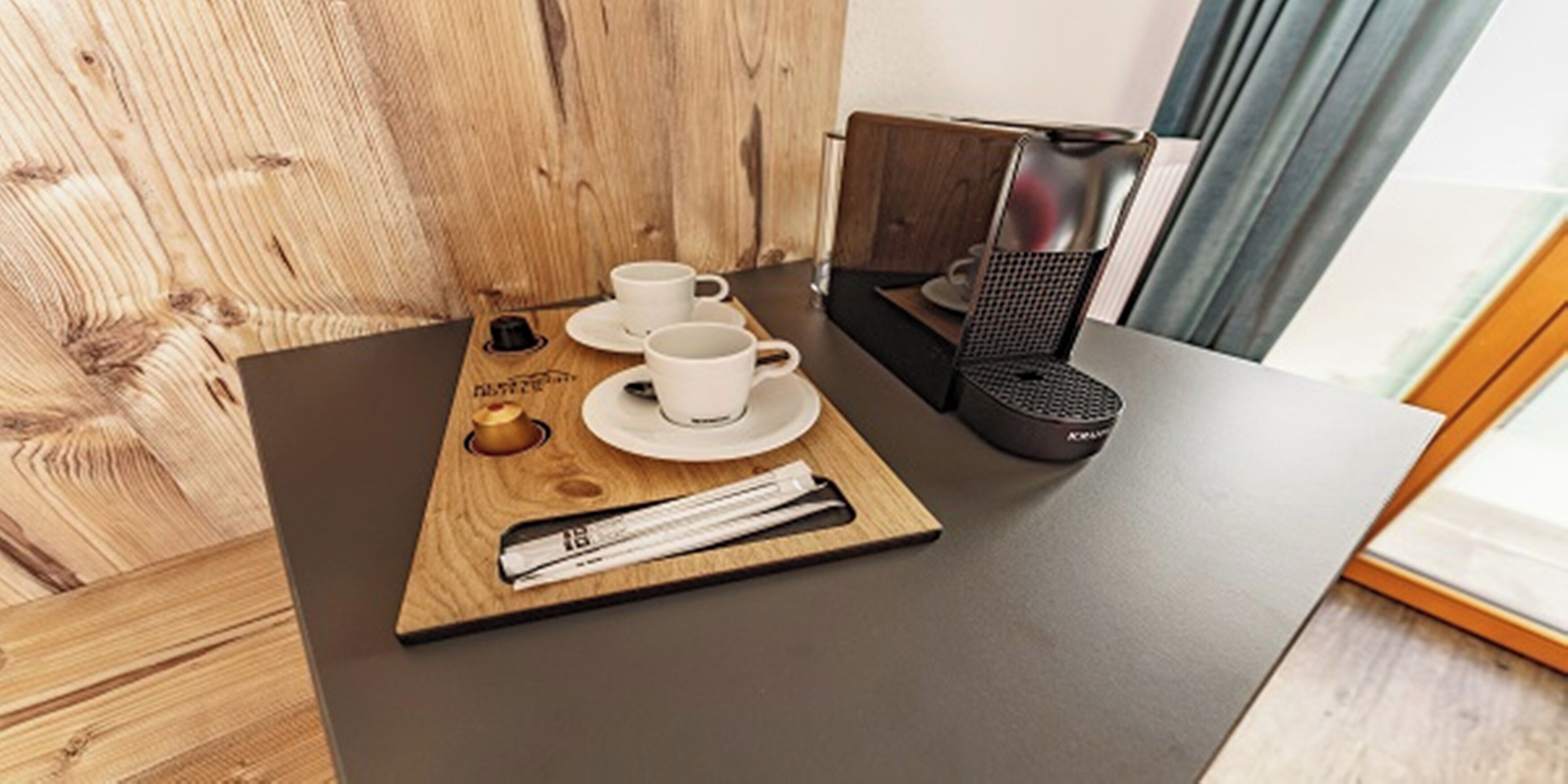 A wooden table with two white cups, a small plate, a black coffee maker, and a newspaper.