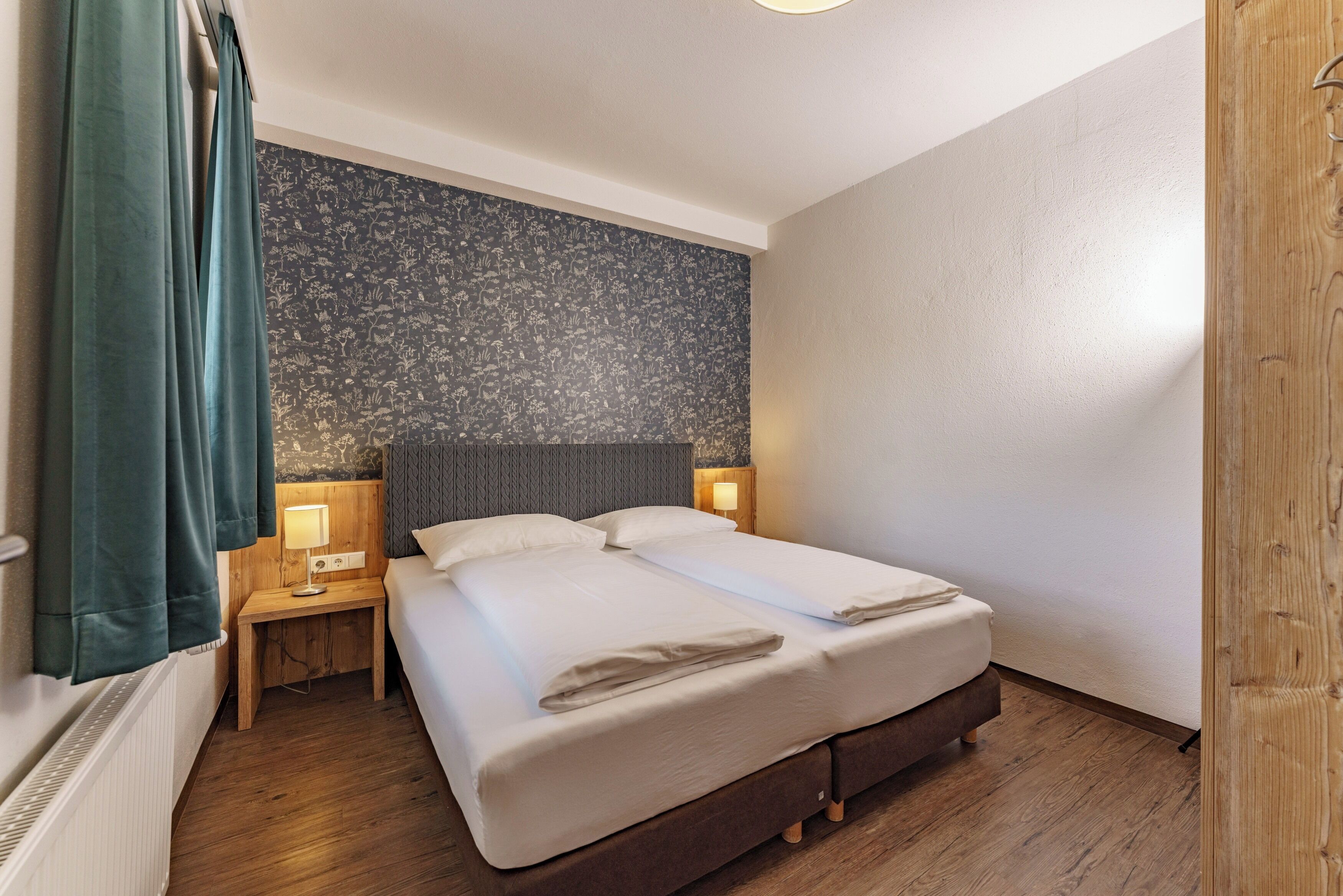 A hotel room with a large bed, two bedside tables, a nightstand, a wooden floor, a patterned wall, and a window with curtains.