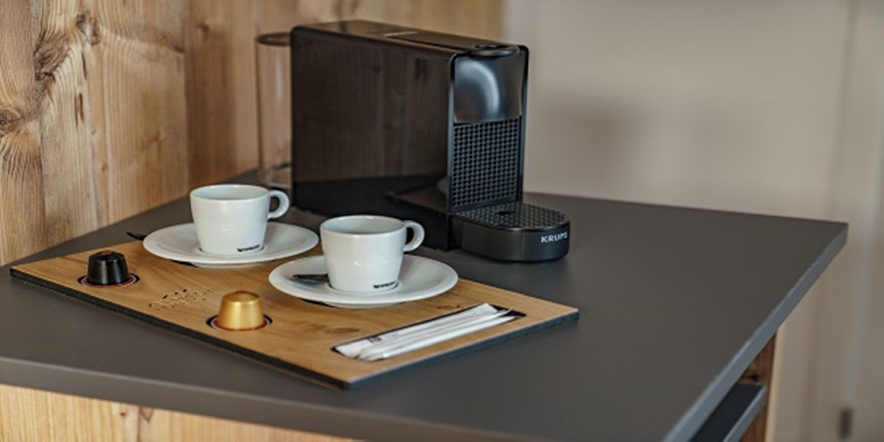 A coffee station with a Krups coffee maker, two white cups, and a wooden tray with a small container and a pen.