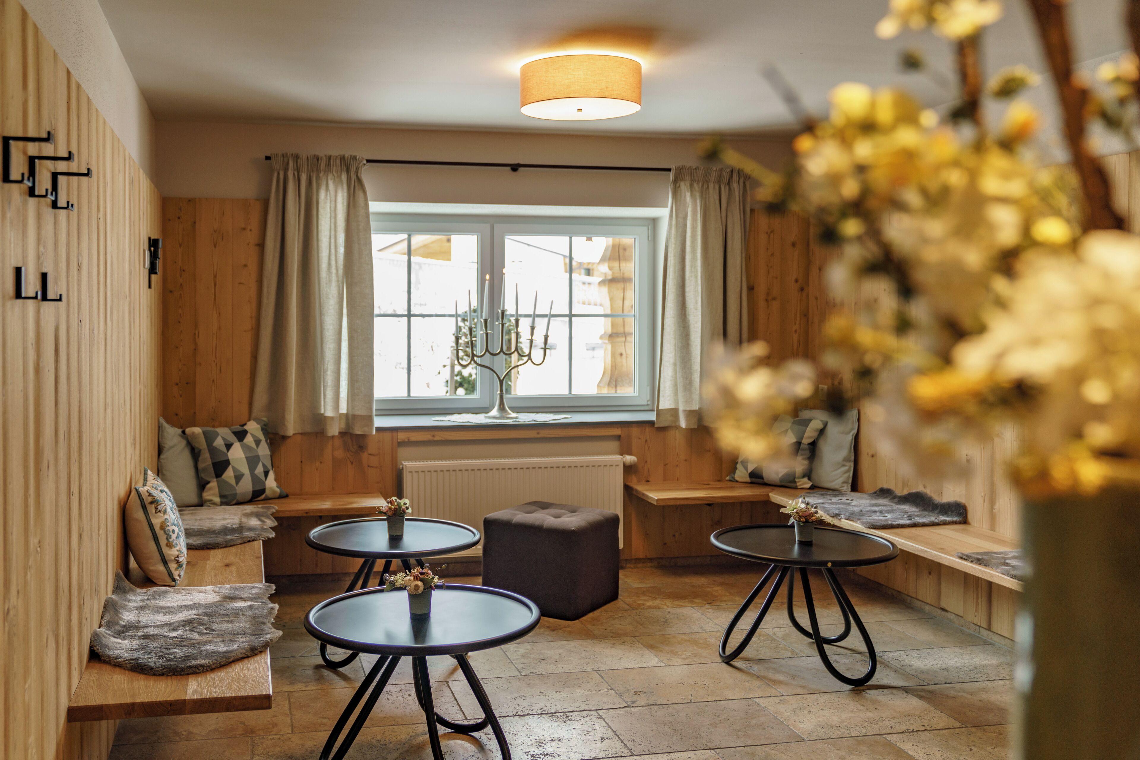 A cozy room with wooden walls, a large window with curtains, a bench, and two round tables with stools.