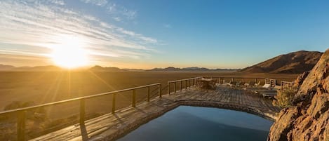 A wooden bridge over a calm body of water, with a sunset in the background and mountains in the distance.