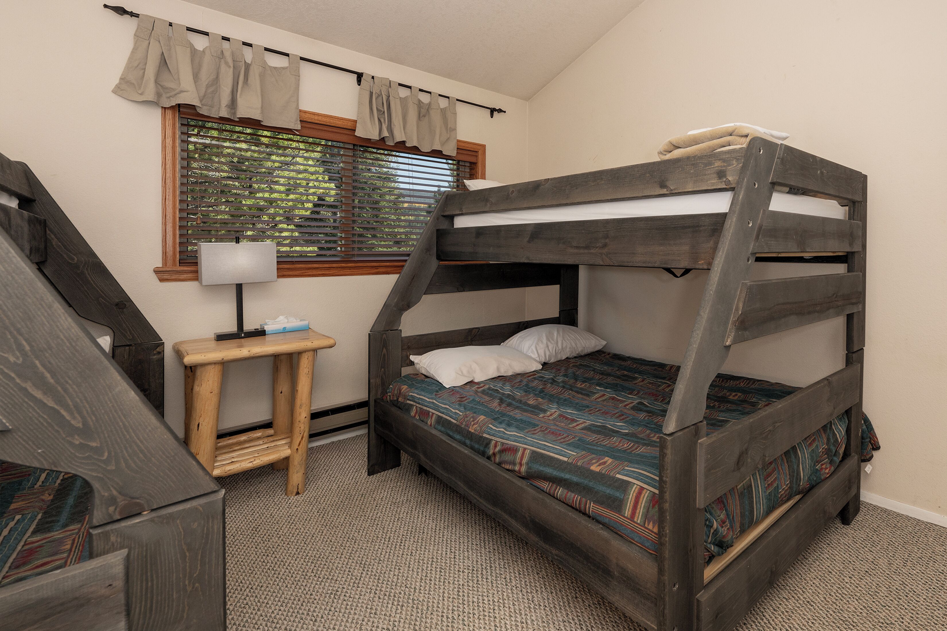 A bunk bed with a wooden frame, a small wooden table with a lamp, and a window with blinds.