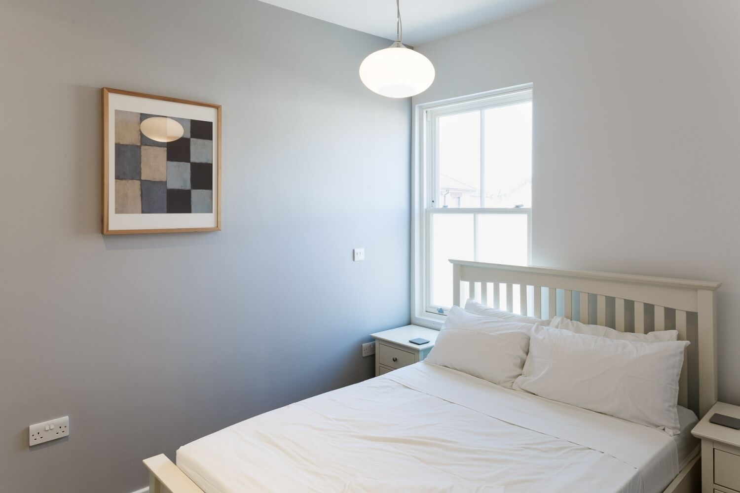 A bedroom with a bed, a nightstand, a bedside table, a window, and a wall-mounted light fixture.