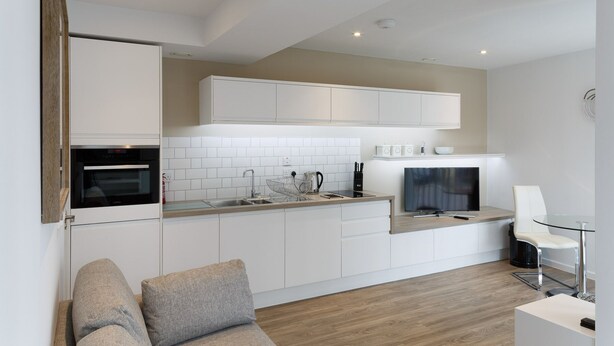 Una cucina moderna con mobili bianchi, forno integrato, lavello e zona pranzo con tavolo in vetro e una sedia.