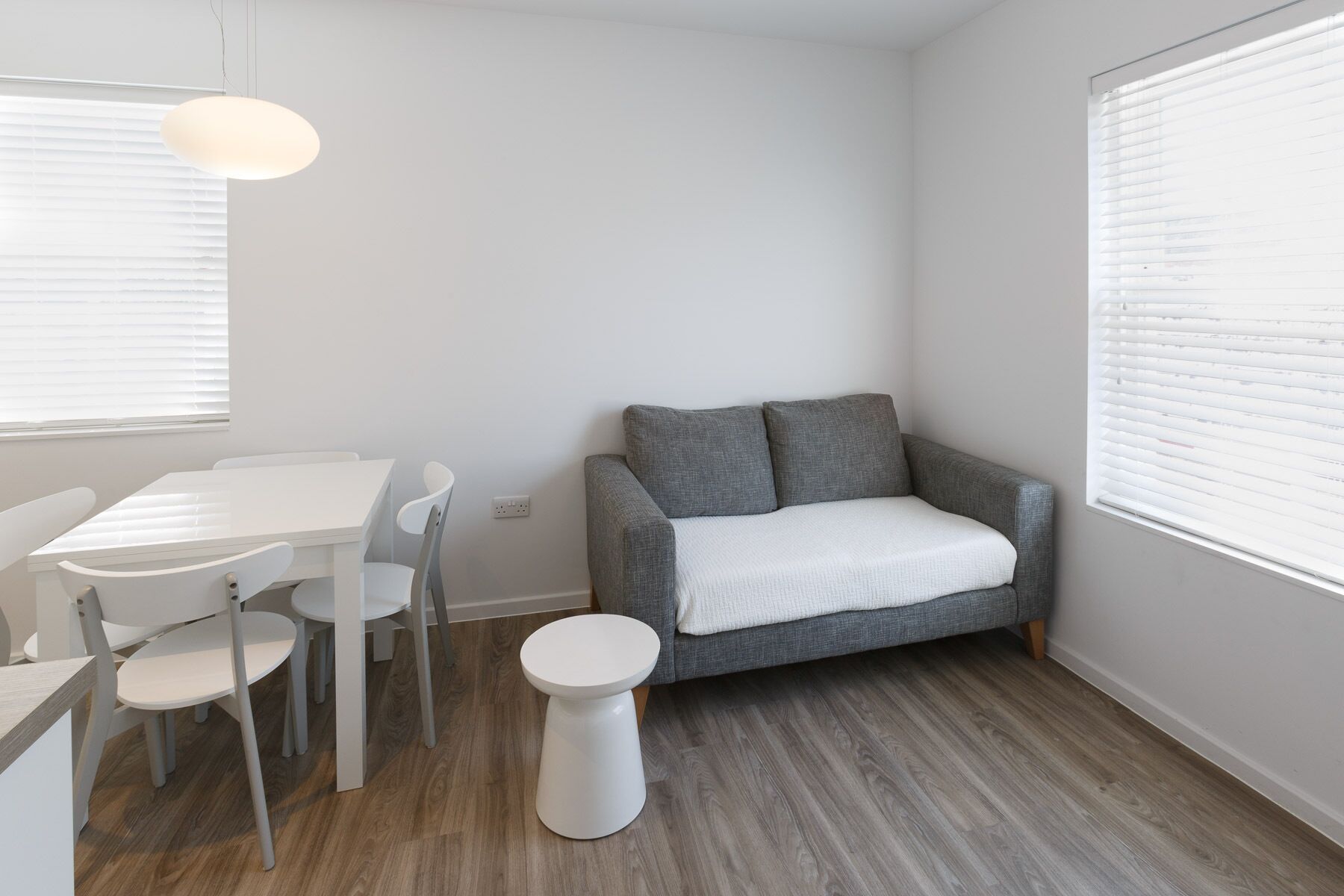 A small, modern room with a grey sofa, a white dining table with chairs, and a small white stool.