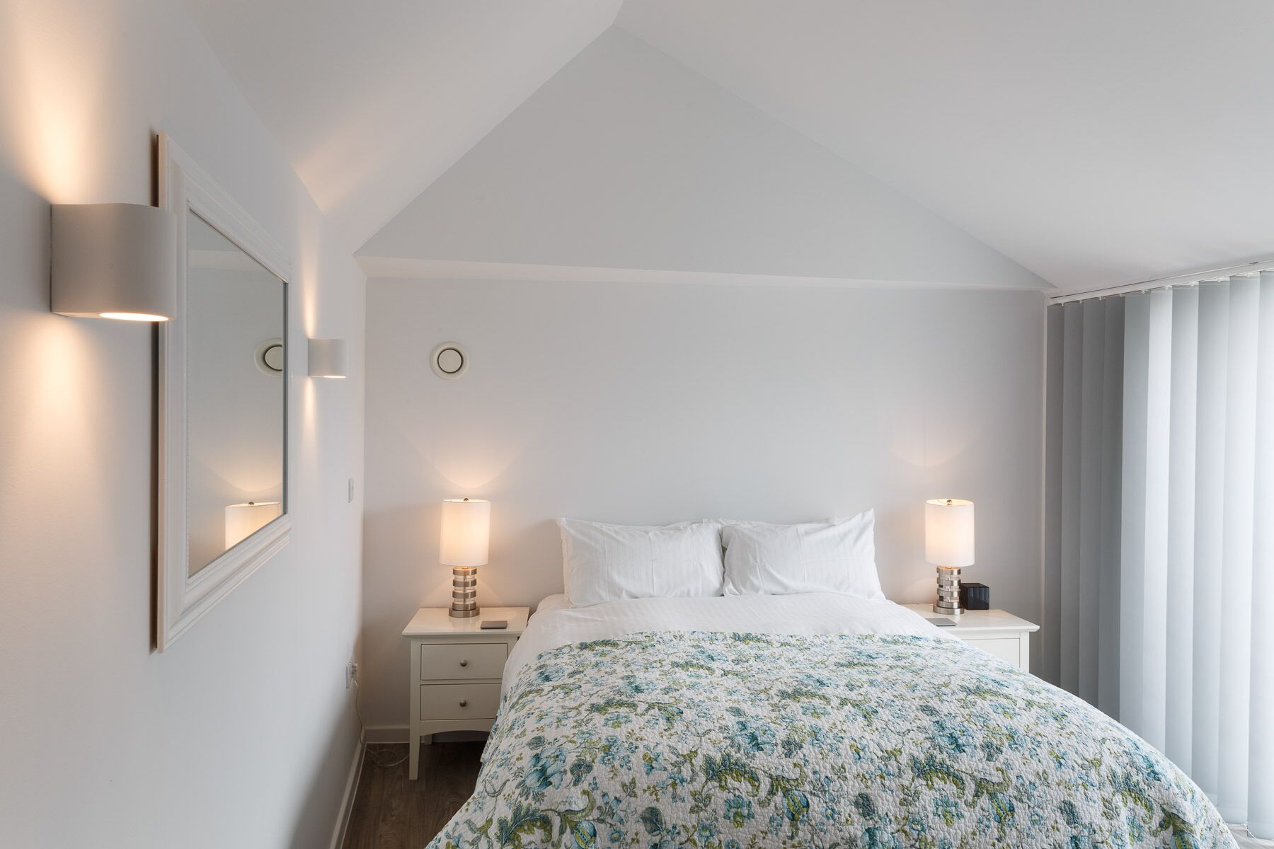 Una habitación de hotel con una cama, dos mesitas de noche, un tocador, un espejo y una ventana con cortinas.