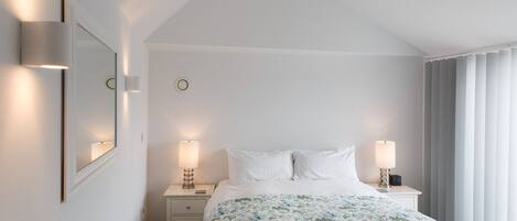 Una habitación de hotel con una cama, dos mesitas de noche, un tocador, un espejo y una ventana con cortinas.