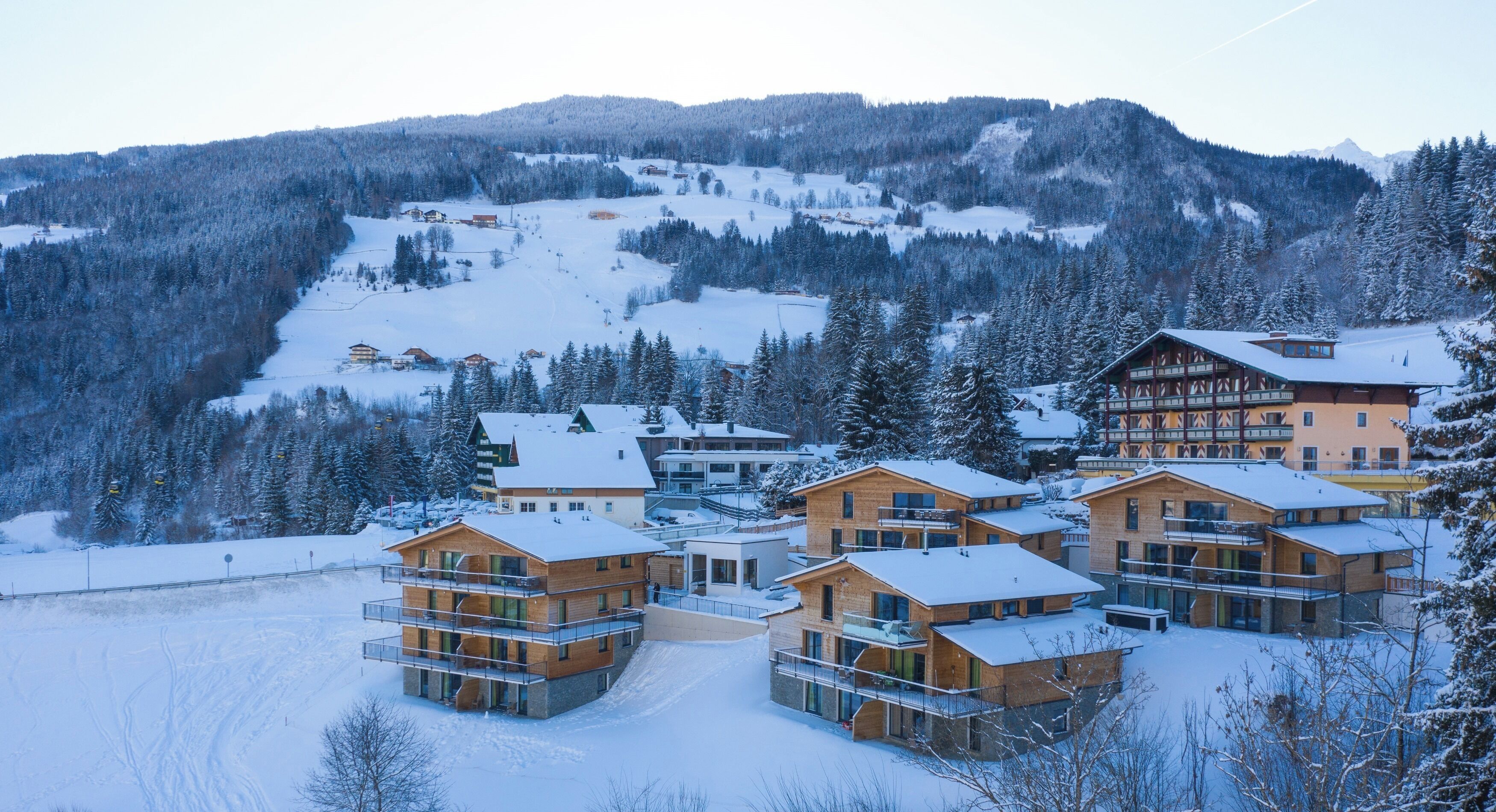 A snow-covered resort with multiple wooden buildings, ski slopes, and a mountainous backdrop.