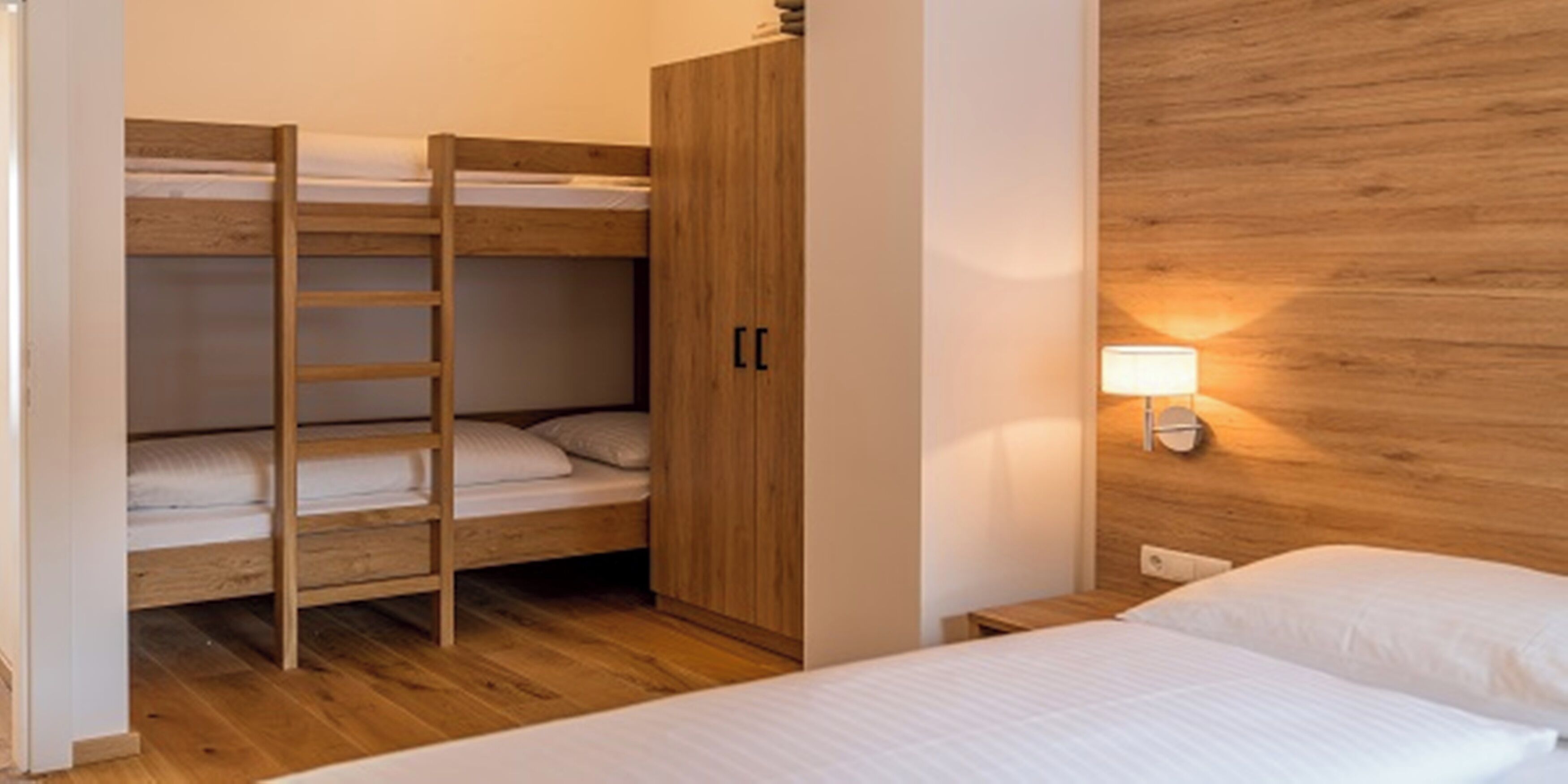 A bunk bed room with a wooden ladder, a wardrobe, and a bedside lamp.