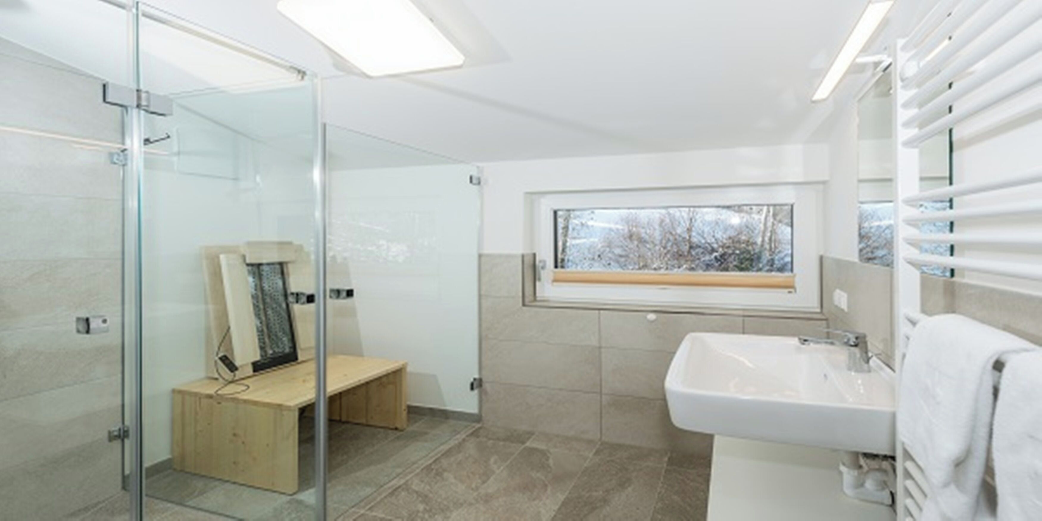 Une salle de bain moderne avec une douche à l’italienne en verre, un banc en bois, une grande fenêtre offrant une belle vue et un lavabo blanc.