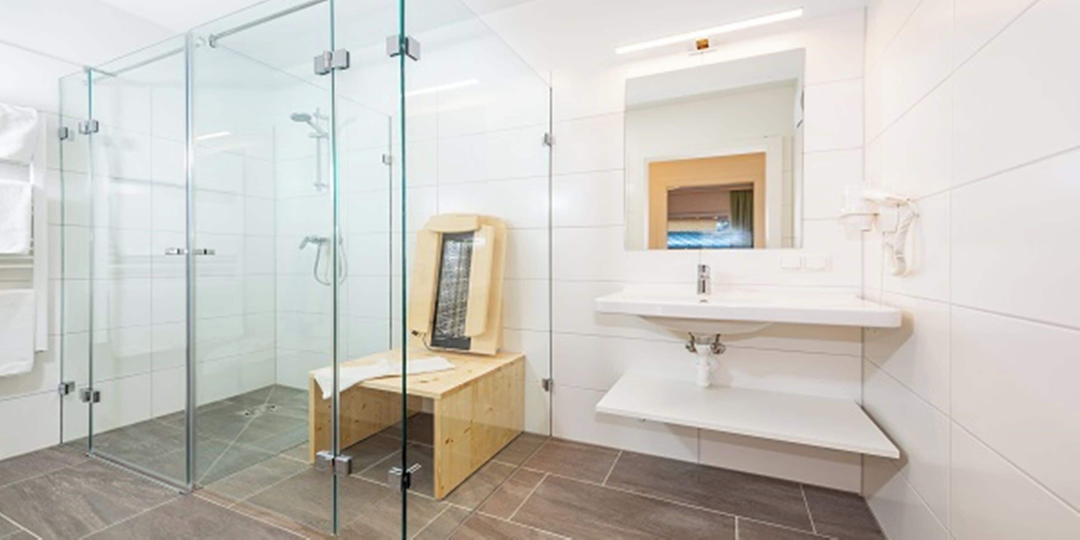Een moderne badkamer met een douchecabine van glas, een houten bankje en een witte wastafel met een spiegel erboven.