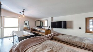 A modern bedroom with a large bed, a bedside table, a mirror, a TV mounted on the wall, and a window with blinds.