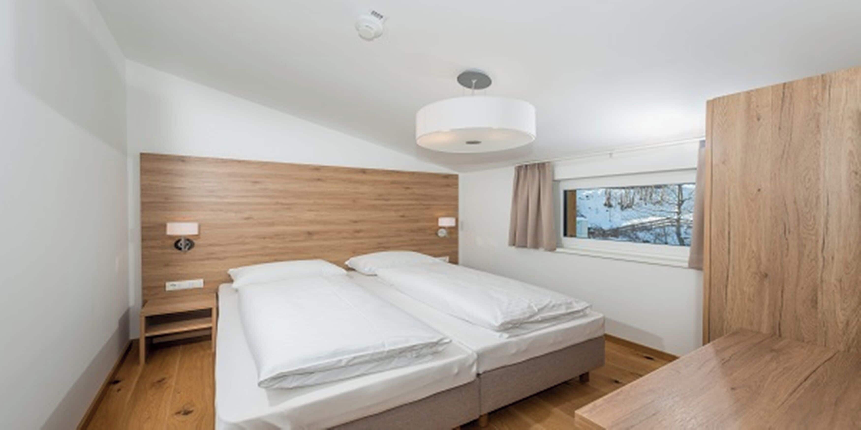 A bedroom with a wooden headboard, two beds, bedside lamps, a window with a view of trees, and a ceiling light.