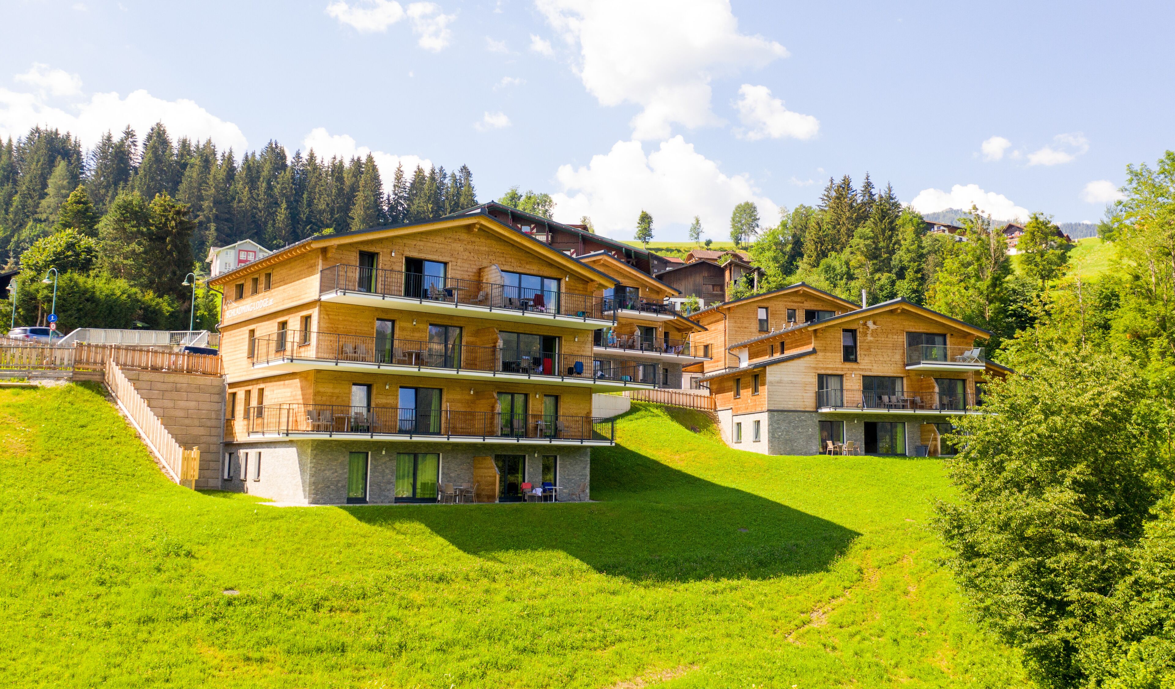 A multi-story wooden building with balconies, set on a grassy hillside with a forested background.