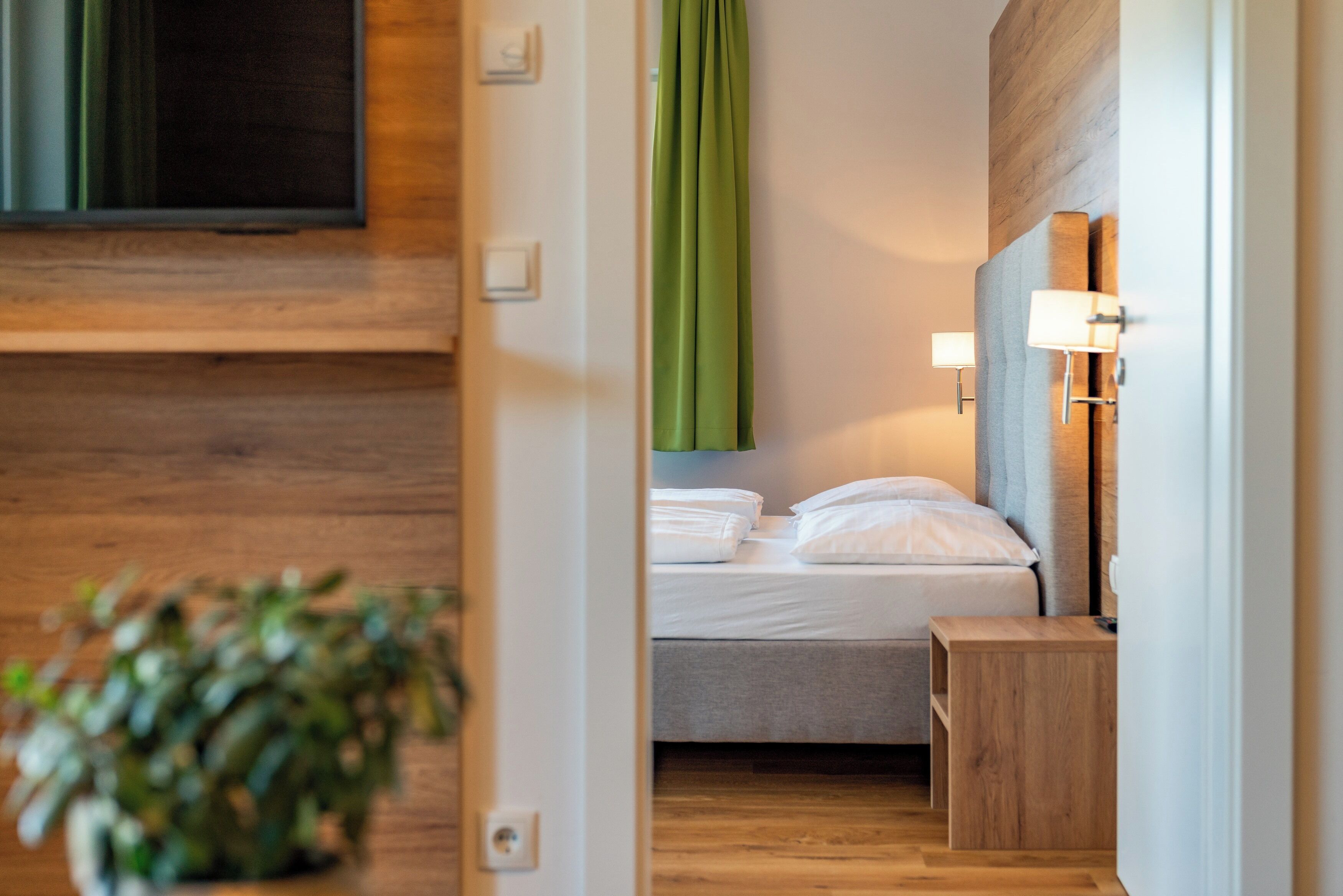 Una habitación de hotel moderna con suelo de madera, una cama con ropa blanca, una mesita de noche y una cortina verde.
