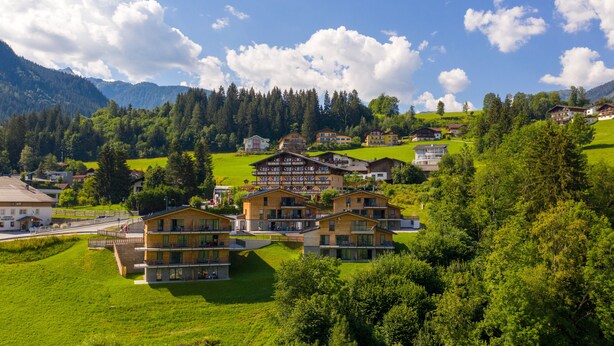 A hillside village with traditional wooden houses, a prominent hotel with a large balcony, and lush green surroundings.