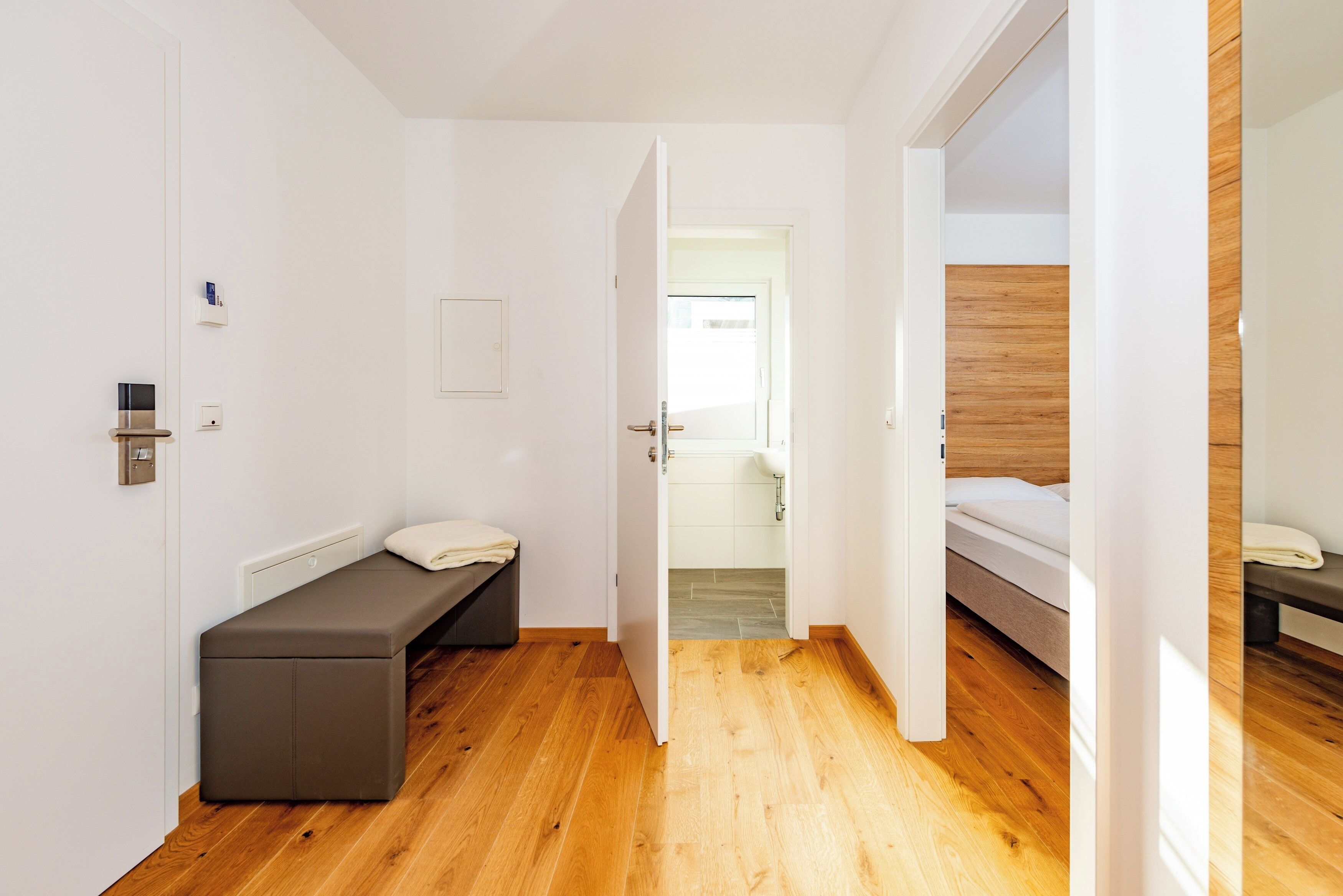Ein modernes Hotelzimmer mit Holzboden, einem Bett, einer Bank und einer Tür zu einem weiteren Raum.