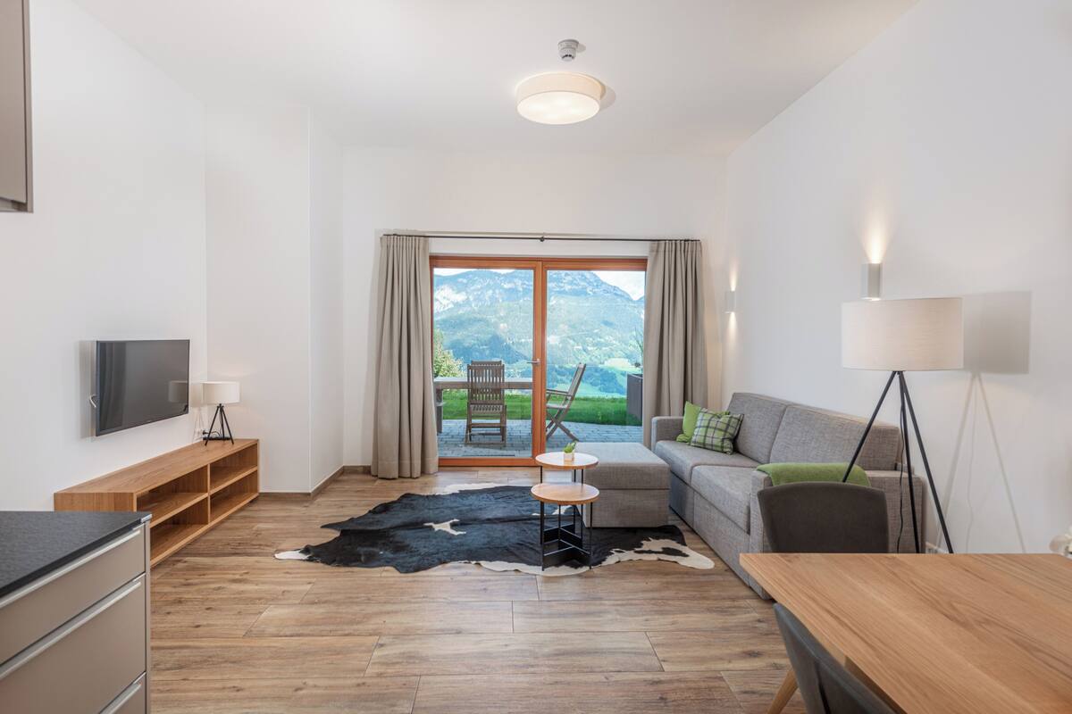 A modern living room with a wooden floor, a sofa, a TV, a small table, and a view of mountains through a glass door.