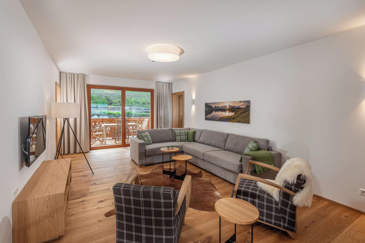 A modern living room with a sectional sofa, a wooden coffee table, and a plaid armchair. There is a large window with curtains, a painting on the wall, and a flat-screen TV mounted on the wall.