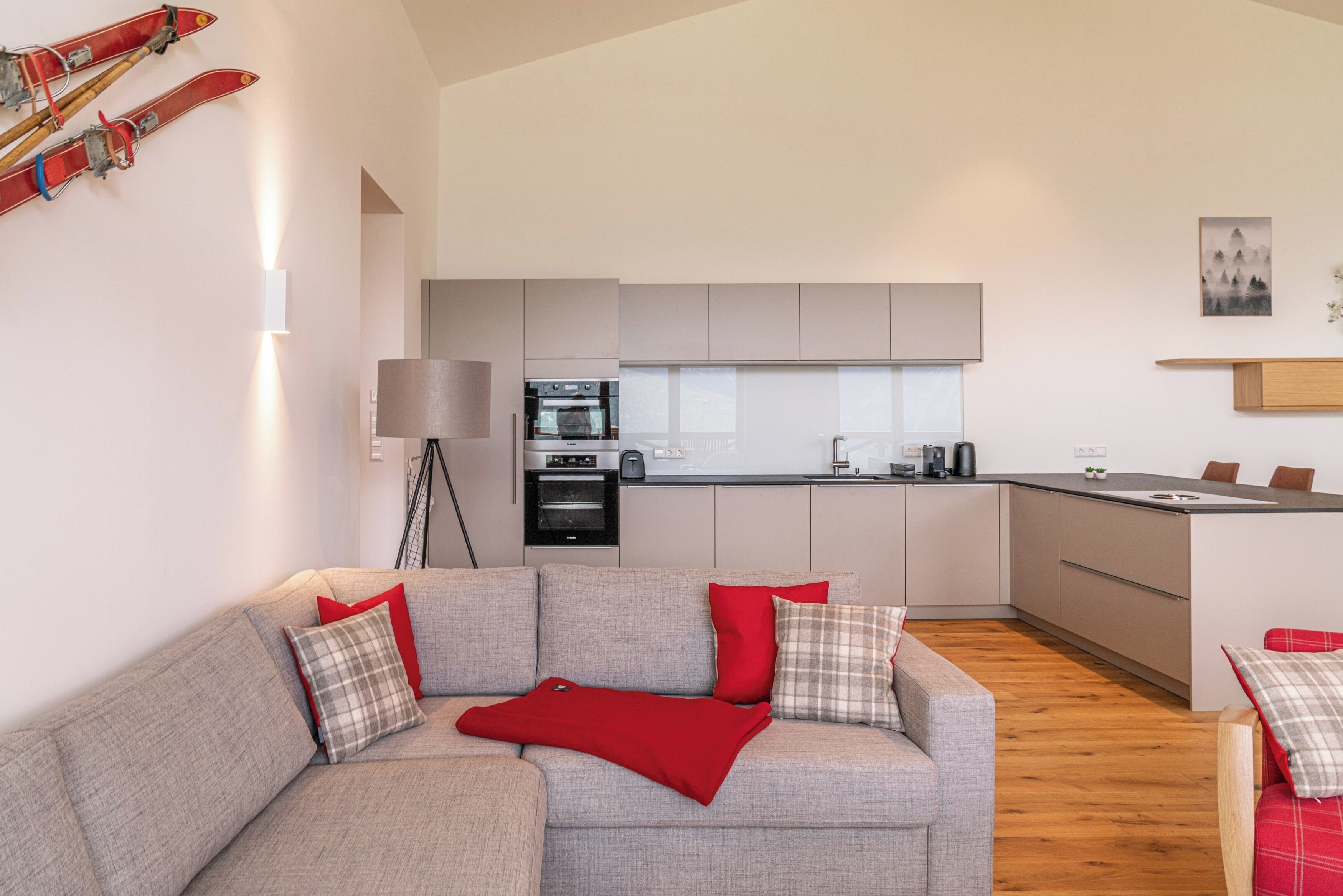 Ein modernes Wohnzimmer mit einer grauen Couch, roten und karierten Kissen, einer roten Decke und einer Lampe. Im Hintergrund ist eine Küche mit weißen Schränken und Geräten zu sehen.