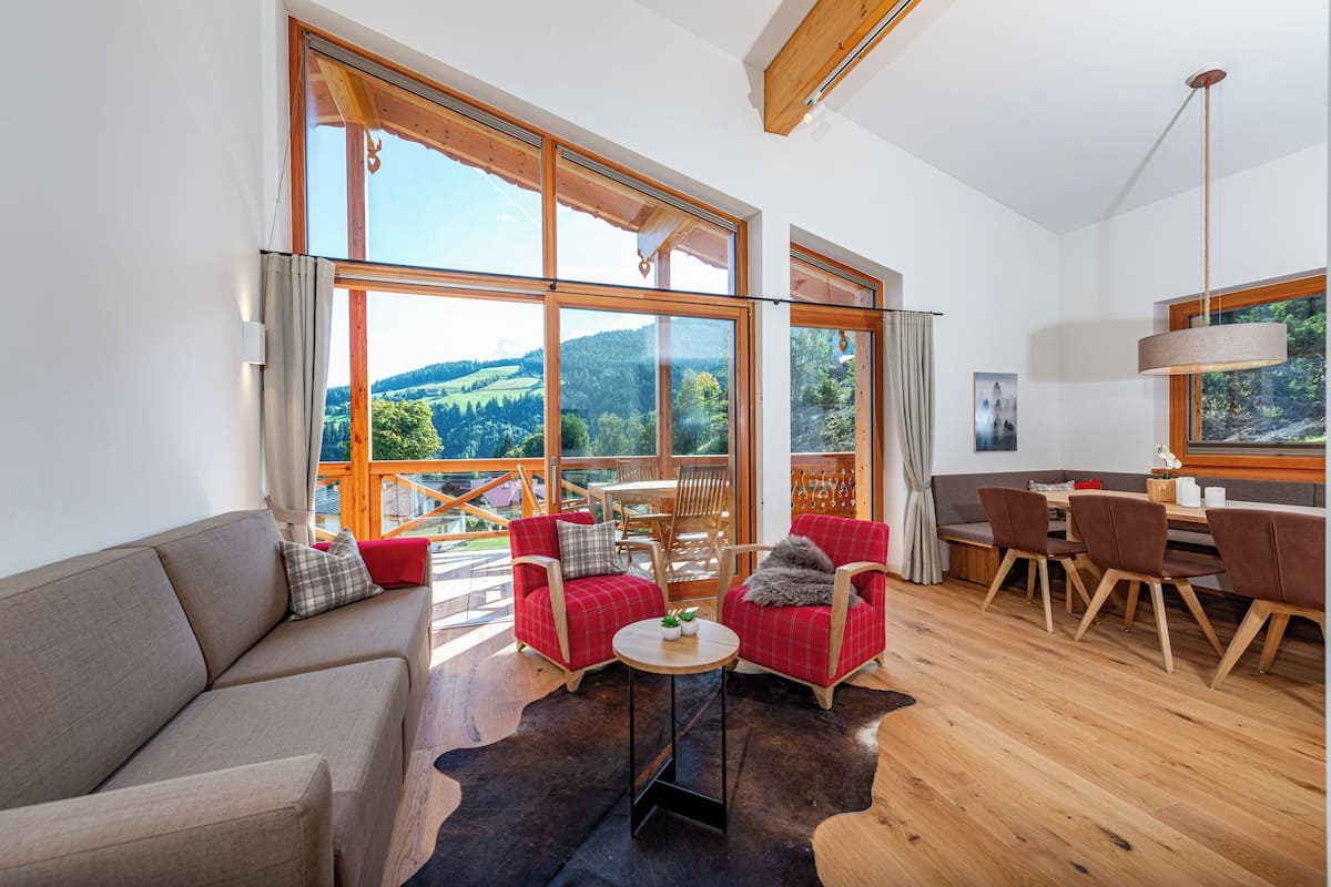 A spacious living area with large windows, a sofa, two armchairs, a round table, and a dining set.