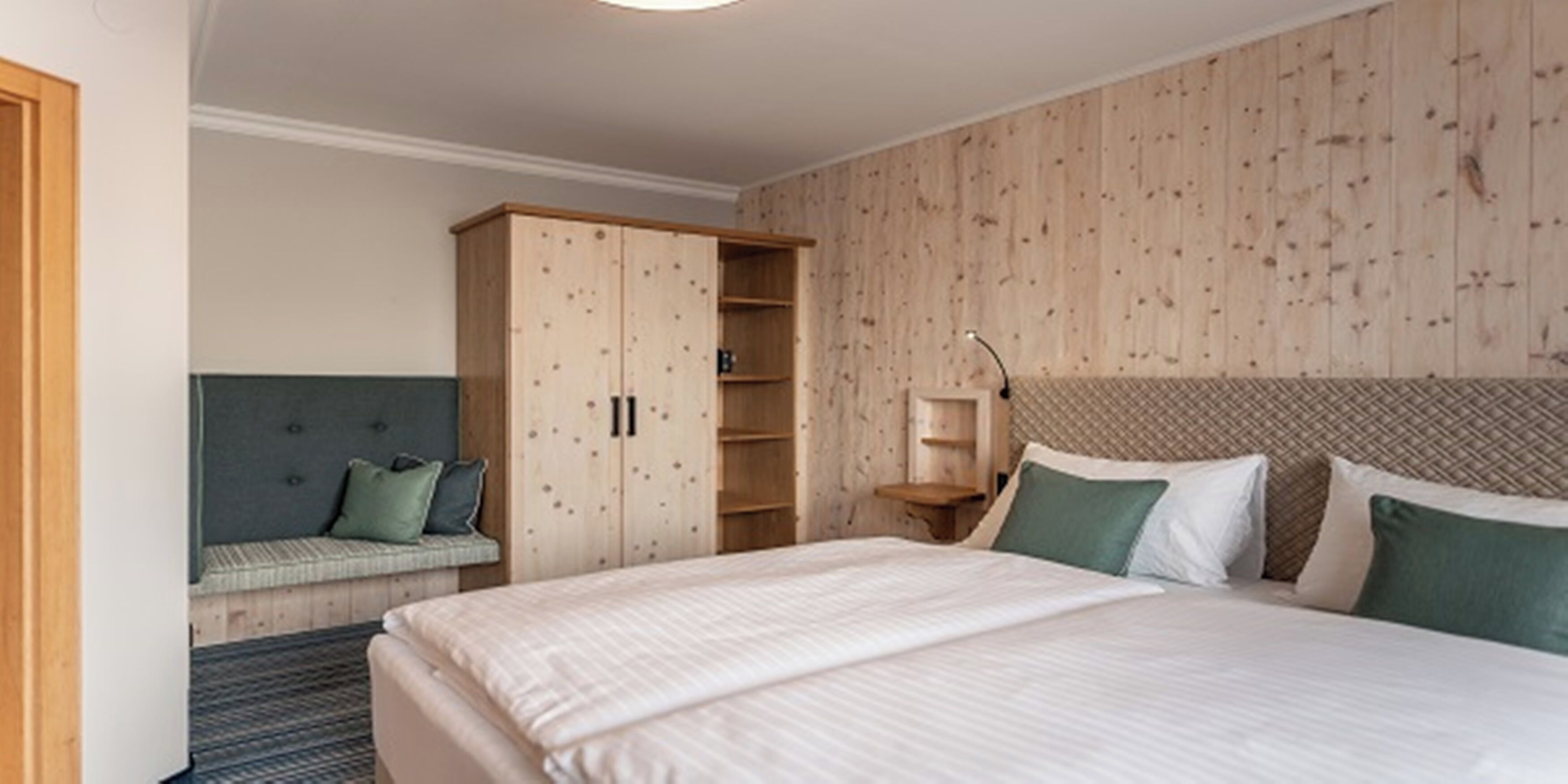 A bedroom with a large bed, a wooden wardrobe, a bedside table, and a bench with cushions.