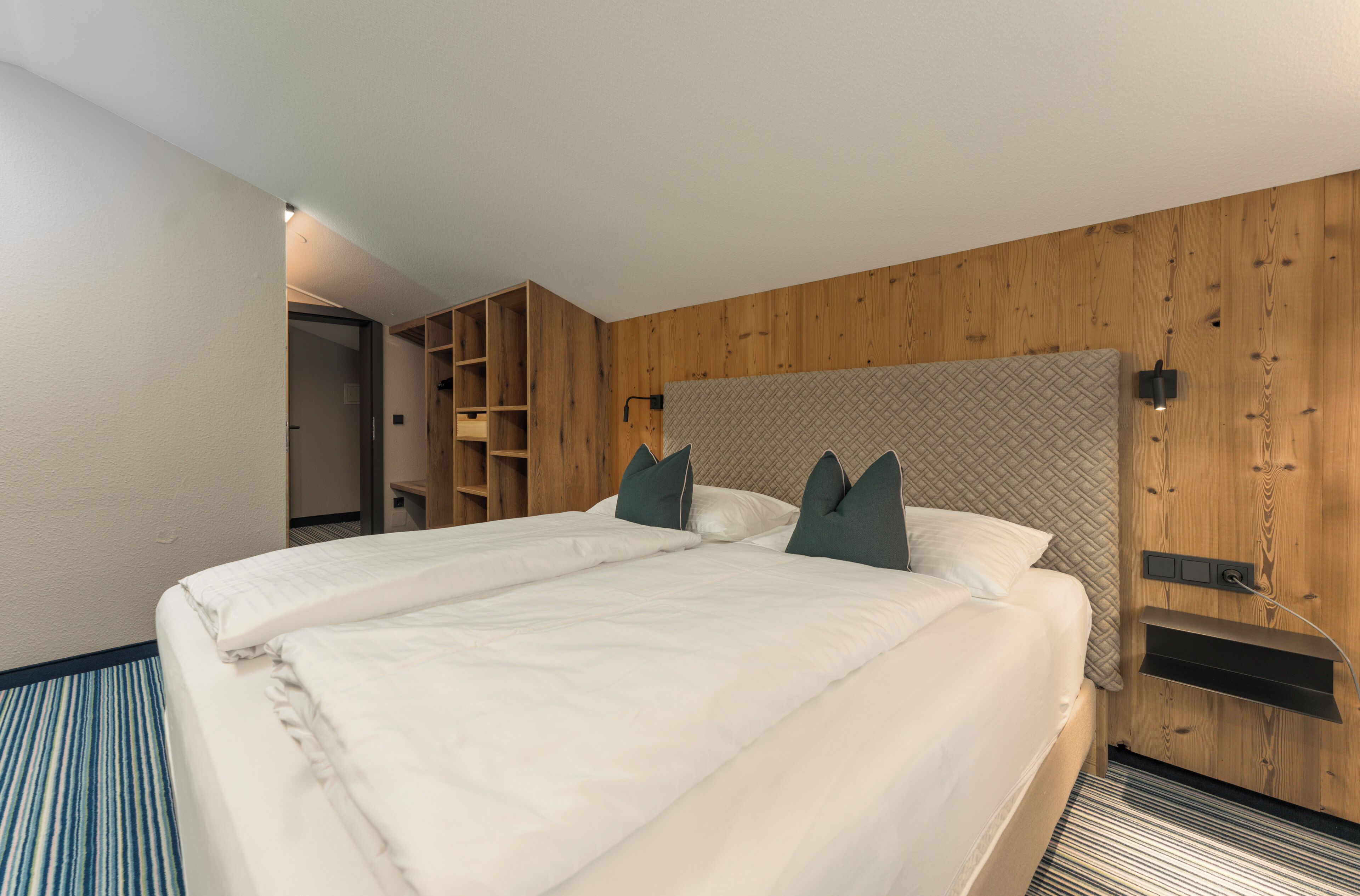 Une chambre d’hôtel moderne avec un grand lit, des lambris en bois aux murs, une étagère intégrée et une moquette à rayures.