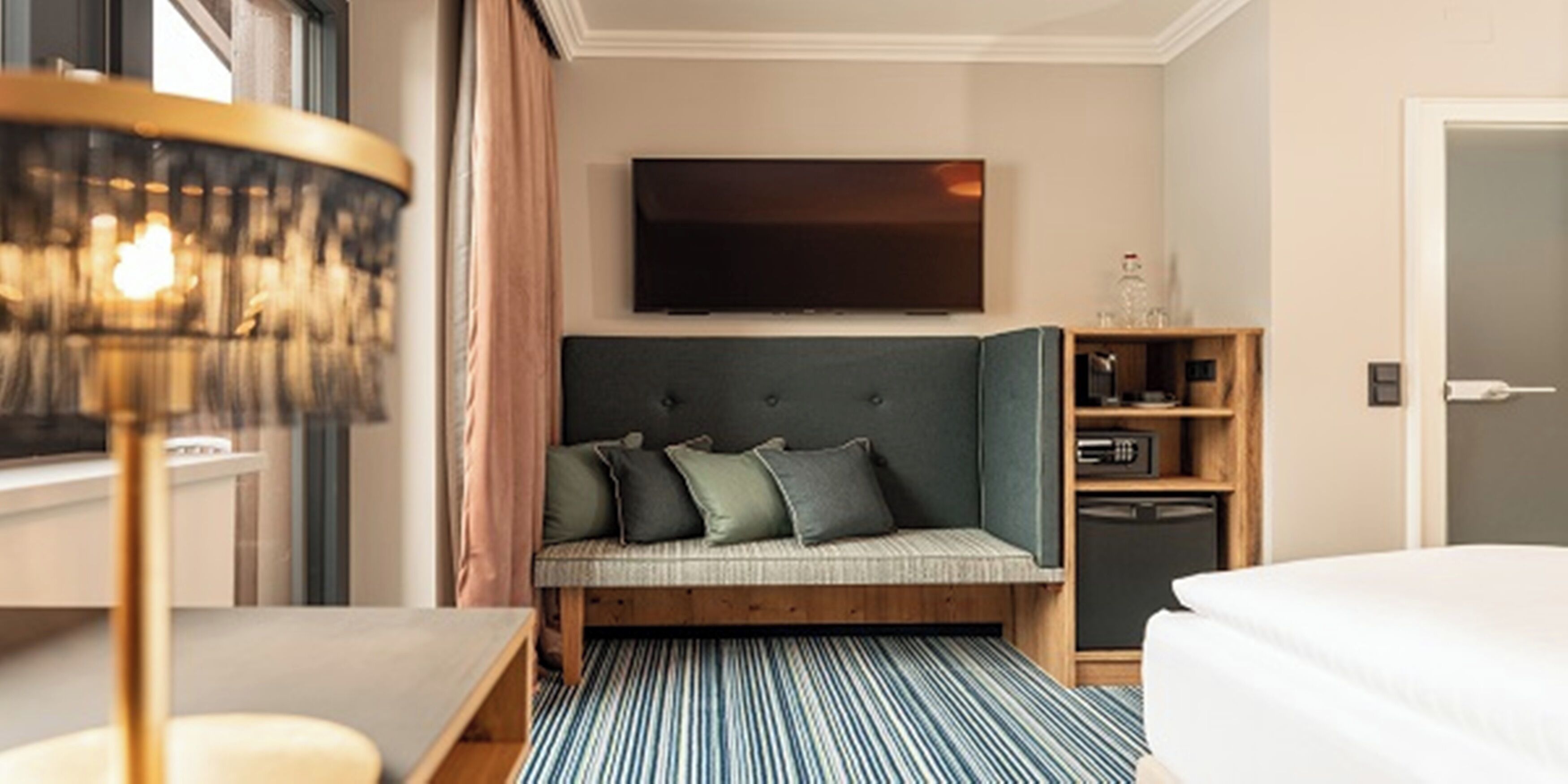 A hotel room with a bed, a sofa bed with cushions, a television mounted on the wall, a small wooden cabinet, and a striped rug.