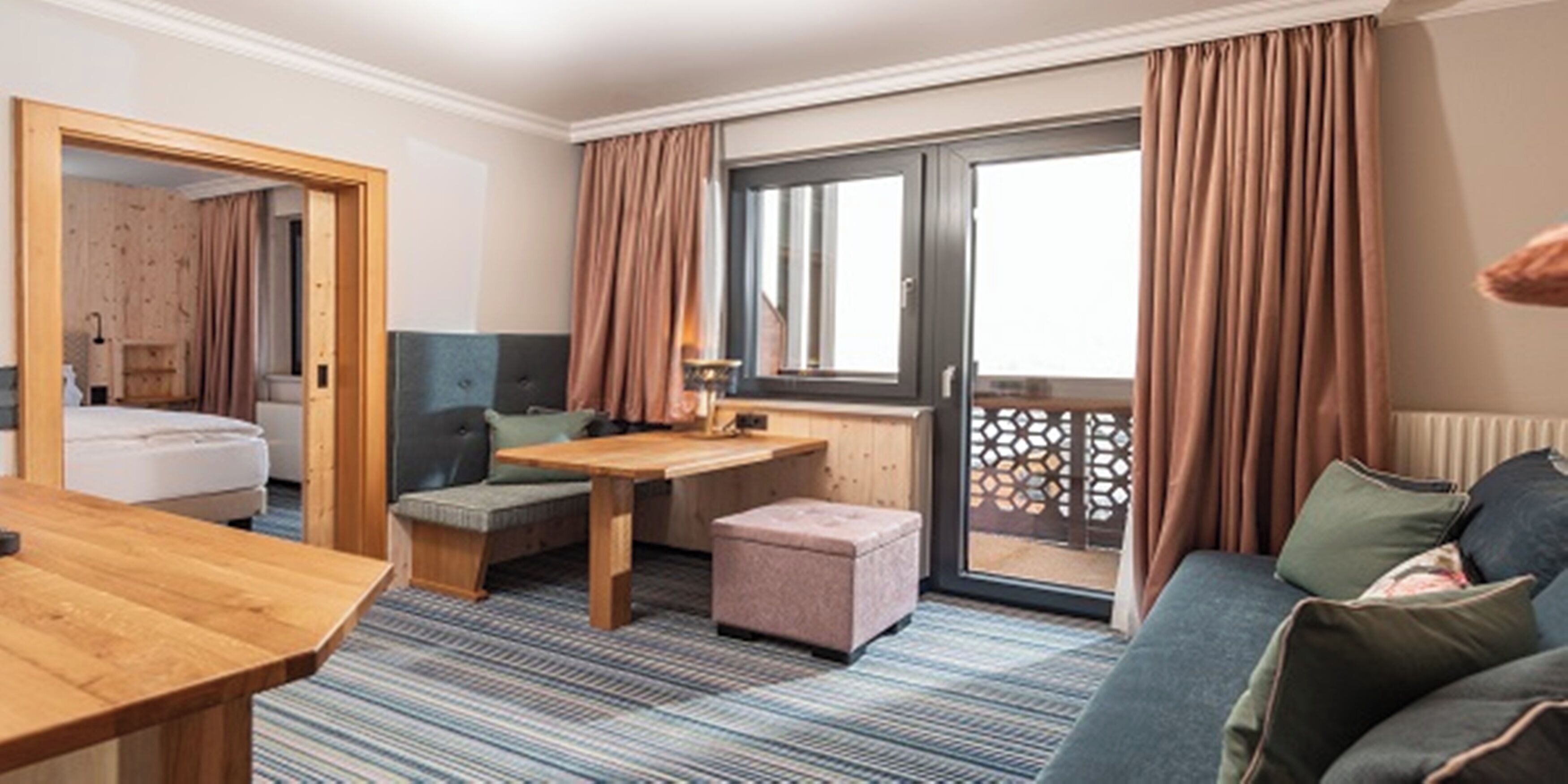 Une chambre d’hôtel avec un lit, une table en bois, un canapé, un petit pouf et une vue sur l’extérieur par une fenêtre avec des rideaux.