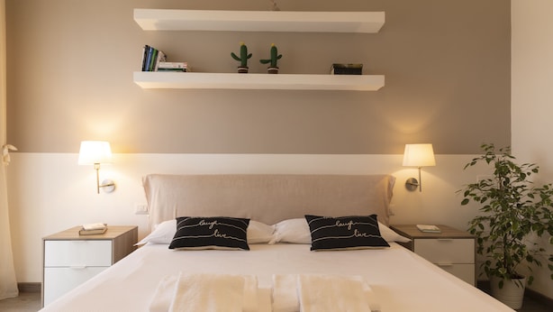 Quarto de hotel bem arrumado com uma cama grande, duas mesas de cabeceira, um criado-mudo, uma prateleira fixada na parede com livros e cactos, e uma planta em vaso.