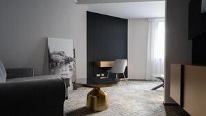 Quarto de hotel moderno com design minimalista, apresentando uma mesa redonda com base dourada, um sofá cinza, uma obra de arte em preto e branco na parede e uma parede de destaque escura.