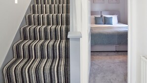 A staircase with striped carpet leading to a bedroom with a bed, a nightstand, and a wall-mounted light fixture. - Newly Refurbished 4 Double Bed House with Parking (Slough)