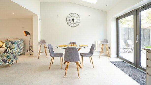 A modern dining area with a round table, grey chairs, and a wall-mounted clock. - Newly Refurbished 4 Double Bed House with Parking (Slough)
