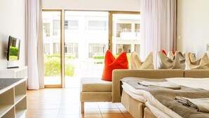 A modern hotel room with a bed, a sofa, a TV, and a balcony with a view of the building and greenery. - Sunwing Sandy Bay Beach (Ayia Napa)