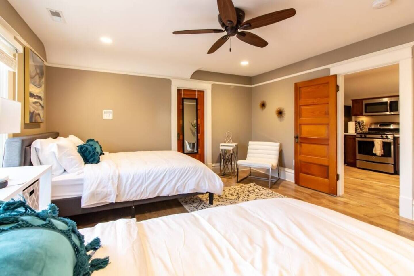 Une chambre avec deux lits, un ventilateur de plafond, une petite table, une chaise et une porte en bois donnant sur une cuisine.