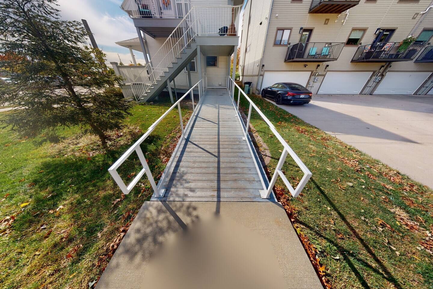 A modern apartment building with a central staircase, a car parked in the driveway, and a tree to the left.